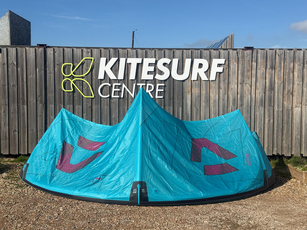 A turquoise kiteboarding kite with magenta graphics set up on gravel in front of a wooden fence with a 'KITESURF CENTRE' sign.