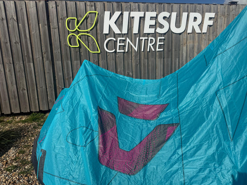 A turquoise blue kitesurfing sail with a purple and gray V-shaped logo, draped in front of a wooden fence featuring the 'Kitesurf Centre' sign.