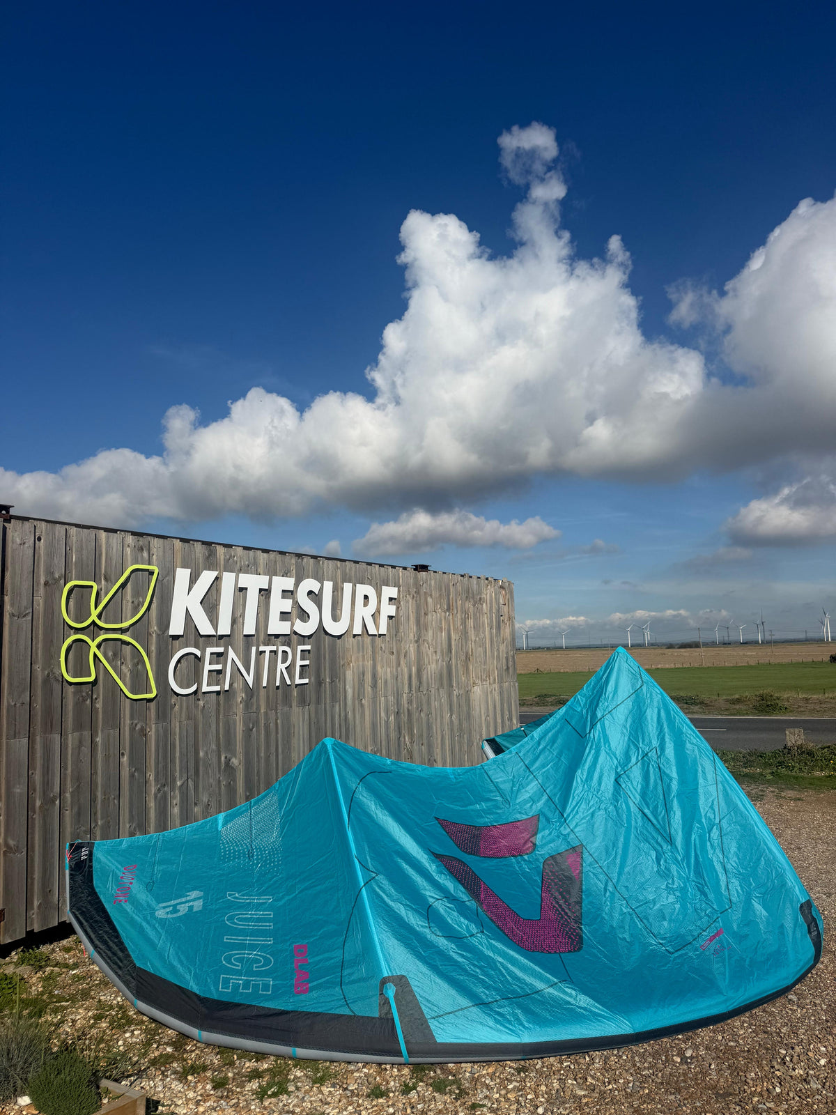 A bright turquoise kiteboarding sail pitched outdoors in front of a wooden Kitesurf Centre sign, with blue sky, white clouds, and open land in the background.