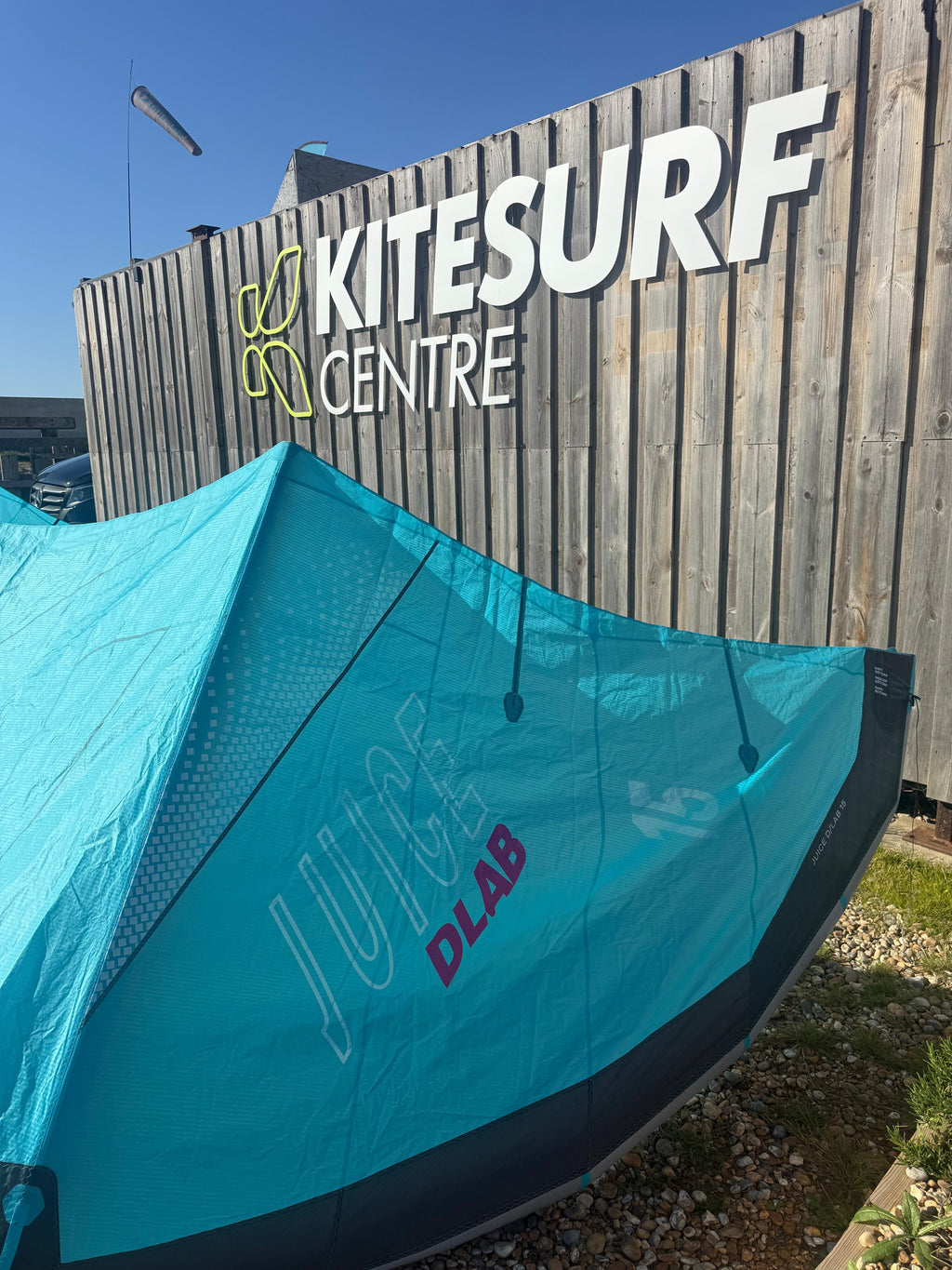 A turquoise and black duotone juice kite  in front of a wooden Kitesurf Centre sign, photographed outdoors on a sunny day.