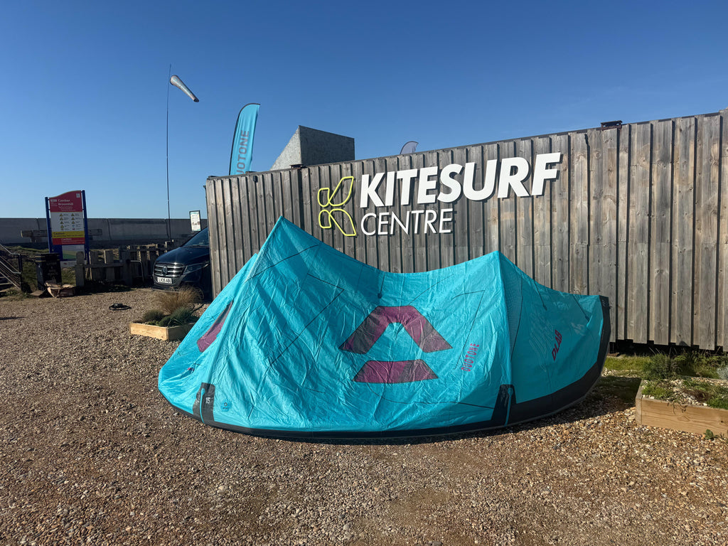 A turquoise kite pitched outdoors in front of a wooden Kitesurf Centre sign under a clear blue sky.