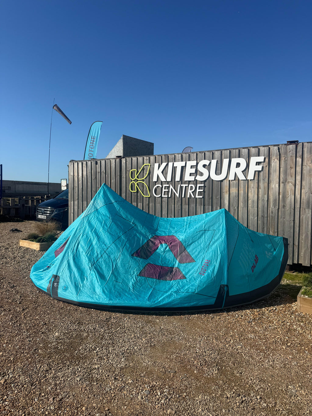 A turquoise kite pitched outdoors in front of a wooden Kitesurf Centre sign under a clear blue sky.